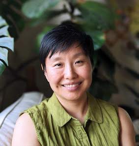 Photo of an Asian woman with short black hair wearing a green sleeveless top with plants in the background